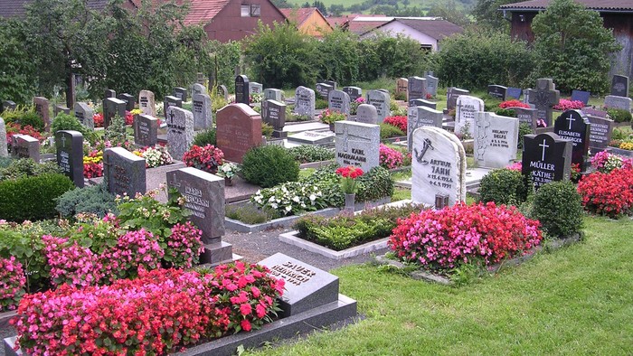 Ein gut gepflegter Friedhof mit bunten Blumen und Grabsteinen, umgeben von Häusern und grünen Hügeln.
