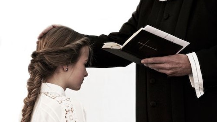 En ung pige med frisyr og hvid blouse ser opad mod en person i svart kapel.