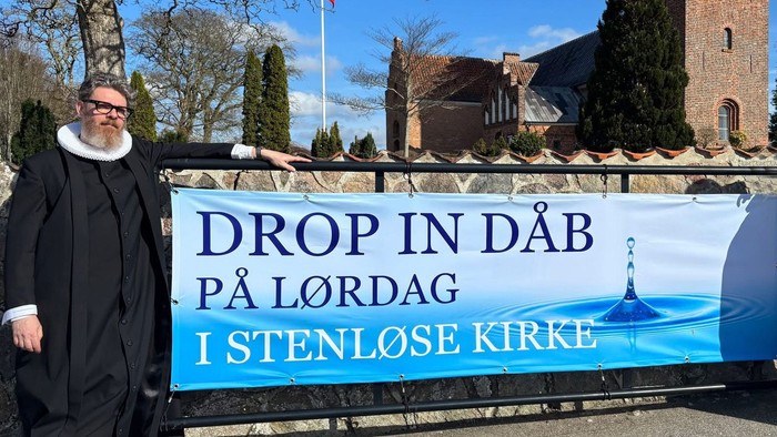 En person i kjortel står ved siden af et banner, der inviterer til dåb i Stensløse Kirke på lørdag.