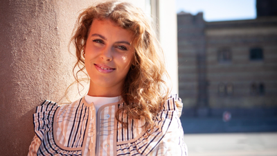 En glad ung kvinde med krøllet hår smiler ved siden af en tekstureret væg i klart dagslys.