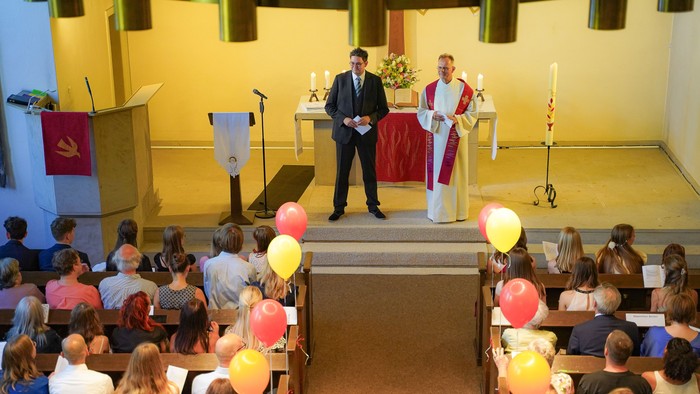 Eine religiöse Zeremonie zeigt einen Priester und einen Zeremonienleiter, die zu einer Gemeinde sprechen, während bunte Luftballons in einer Kirche zu sehen sind.