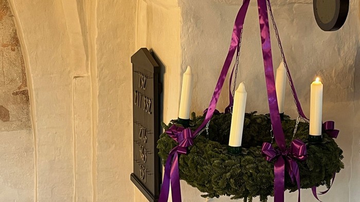En adventskrans med et lys tændt og hængt op i lilla bånd.