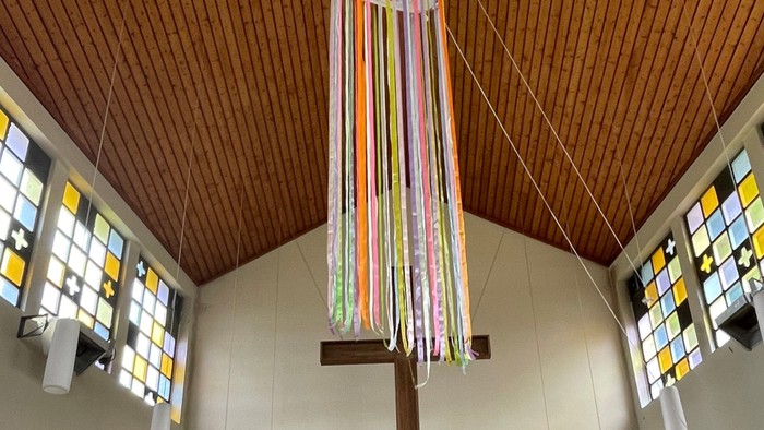 Eleganter Kapelleninnenraum mit einem hohen Kreuz, das mit bunten Bändern geschmückt ist, und farbigen Kirchenfenstern.