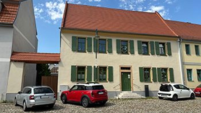 Vor einem gelben Haus mit grünen Fensterläden stehen drei Autos auf Kopfsteinpflaster.