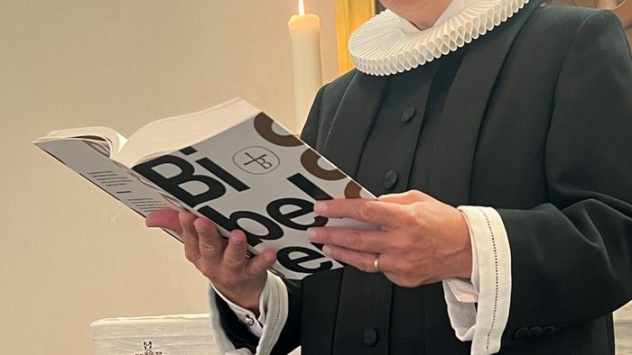 En person i mørkt tøj og hvid krage læser fra en bog med titlen 