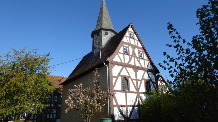 Das Bild zeigt eine Kirche mit einem spitzen Turm und Holzfassade, umgeben von Bäumen und Büschen.