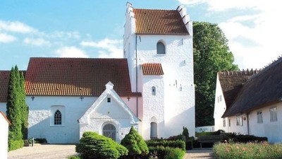 Et hvidt kirkebygningskompleks med røde tag og en tårn, omgivet af grønne buske og træer