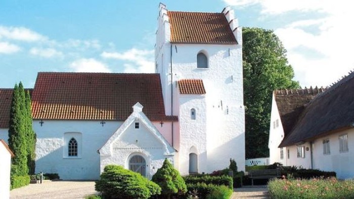 Et hvidt kirkebygningskompleks med røde tag og en tårn, omgivet af grønne buske og træer