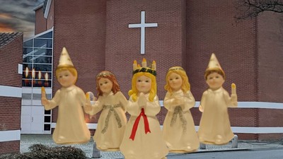 Fem dukker i hvide kjoler og hatte står foran en murstenskirke med et kors.