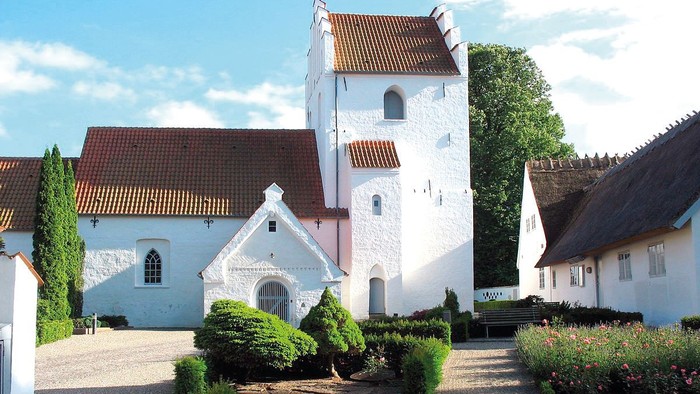 En smuk og hvid kirke med tårn og røde tag i en trædit og grønne omgivelser