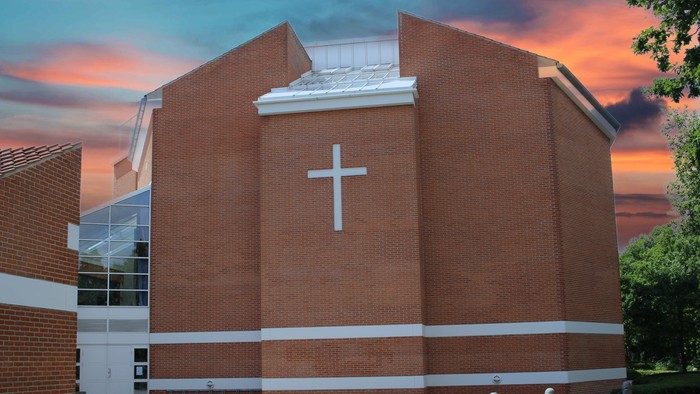 En rød murstenskirke med et stort hvidt kors på fasaden, beliggende ved en vej med træer og en rød skygge.