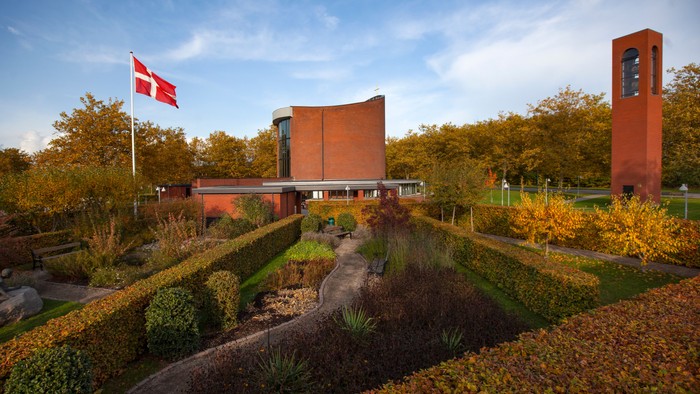 En moderne bygning med en dansk flag i have, omgivet af en park med høje buske og en klokketårn til højre
