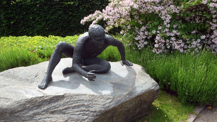 En bronsstatue af en siddende figur på en stor sten i en have med grønne planter og blomster.