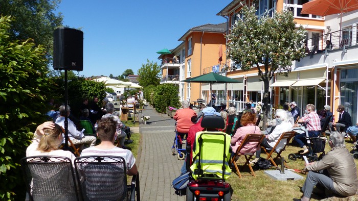 Menschen sitzen auf Bänken und auf dem Rasen in einem öffentlichen Bereich mit Gebäuden und Bäumen im Hintergrund.
