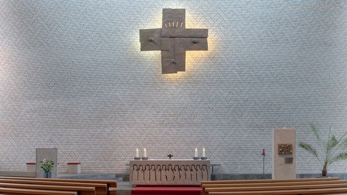 Moderner Altarraum mit Steinaltar, brennenden Kerzen und einem großen beleuchteten Metallkreuz an einer weißen Ziegelwand.