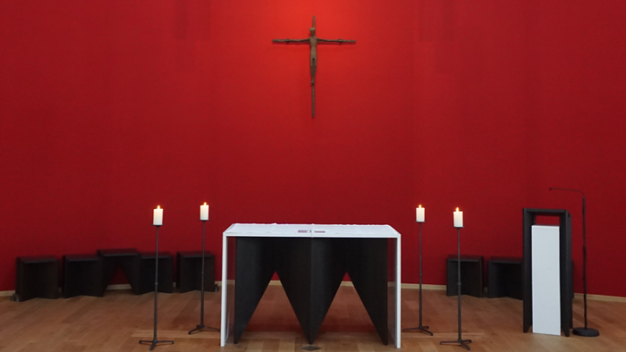 Moderner Altar mit Kruzifix vor einer roten Wand und brennenden Kerzen.