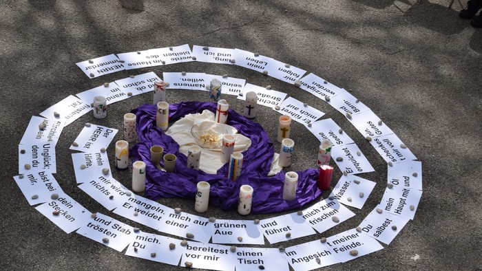 Ein Kreis aus weißen Pappschildern mit deutschen Texten umgibt Kerzen und Blumen auf einem violetten Tuch.