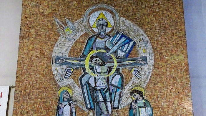 Ein Steinaltar mit Blumenschmuck und zwei Kerzen steht vor einem großen Mosaik-Wandbild. Das Mosaik zeigt zentral die Heilige Dreifaltigkeit.