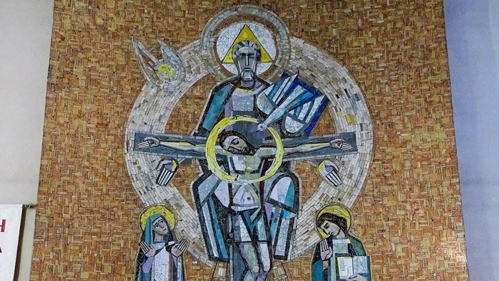 Ein Steinaltar mit Blumenschmuck und zwei Kerzen steht vor einem großen Mosaik-Wandbild. Das Mosaik zeigt zentral die Heilige Dreifaltigkeit.