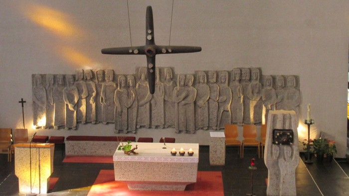 Ein großes Kreuz hängt von der Decke über einem Altar, auf dem drei Kerzen brennen. Links vom Altar befindet sich der Ambo, rechts steht der Tabernakel.