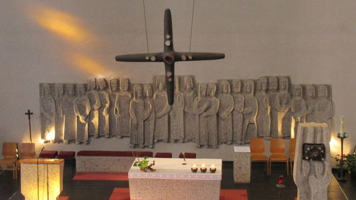 Ein großes Kreuz hängt von der Decke über einem Altar, auf dem drei Kerzen brennen. Links vom Altar befindet sich der Ambo, rechts steht der Tabernakel.