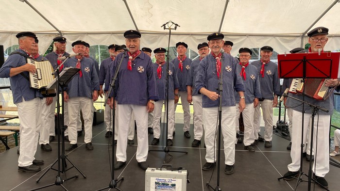 Eine Gruppe von Männern in blauen Uniformen und weißen Hosen singt unter einem Zelt.