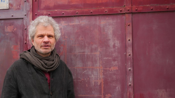 Mann mit grauem Haar vor rotem Metalltor posierend