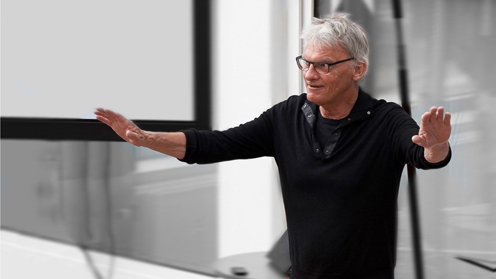 Mand med briller og kort hår står med udstrakt arme foran et hvidt bord.