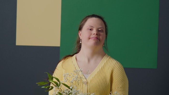 En kvinde i gul trøje står foran en mørk blå vegg med grønne og gule rektangler og en blomst.