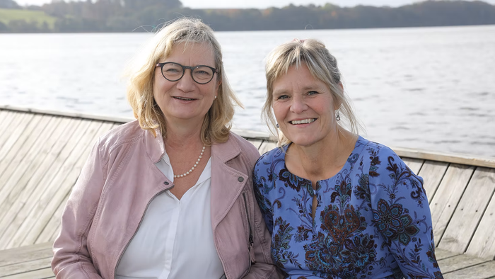 To kvinder sidder ved vandet og smiler til kameraet
