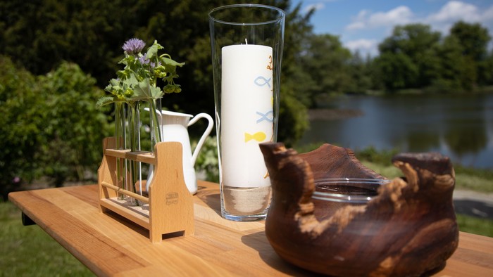 Eine friedvolle Outdoor-Szenerie mit einem Holztisch, auf dem eine Kerze, Blumen und ein geschnitztes Holzobjekt arrangiert sind, mit Blick auf einen ruhigen See.