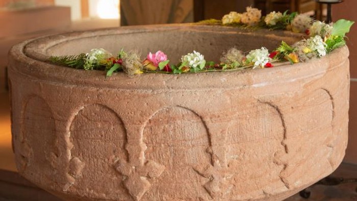 Taufstein, geschmückt mit Blumen; im Hintergrund: Taufkerze und Altar. 