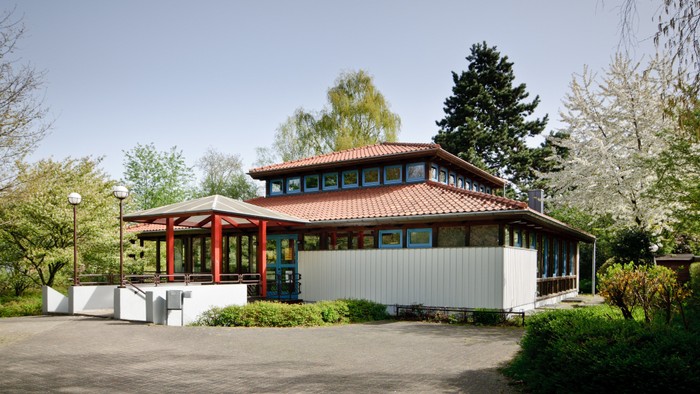 Das Bild zeigt ein modernes Gebäude mit rotem Dach und großen Fenstern, umgeben von Grünflächen und Bäumen.