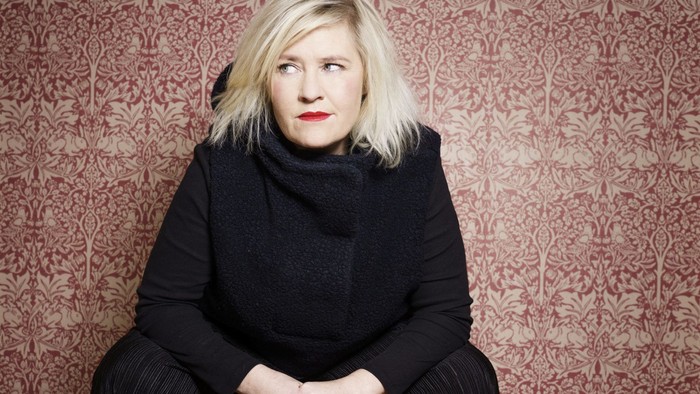 Kvinde med blond hår sidder mod en mønstret rød væg.