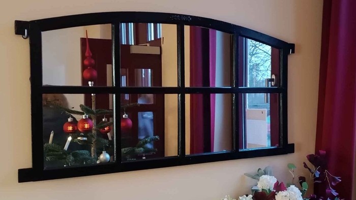 Ein dekorativer Wandspiegel hängt an einer Wand und spiegelt einen Raum mit einem roten Vorhang und einer Vase mit Blumen wider.