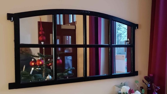 Ein dekorativer Wandspiegel hängt an einer Wand und spiegelt einen Raum mit einem roten Vorhang und einer Vase mit Blumen wider.