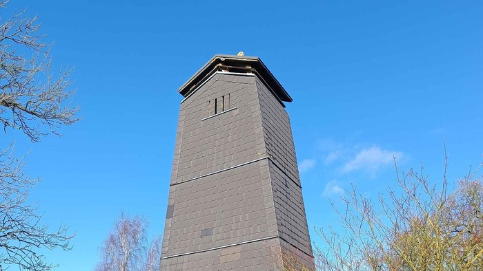 Hoher Steinturm mit Sternemblem und Ziffernblatt in einer gestalteten Parkanlage.