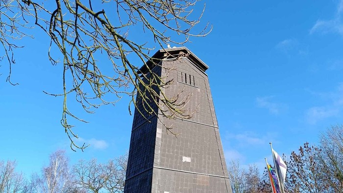 Hoher Uhrturm mit Sternsymbol und klarem blauem Himmel im Hintergrund, umgeben von kahlen Bäumen.