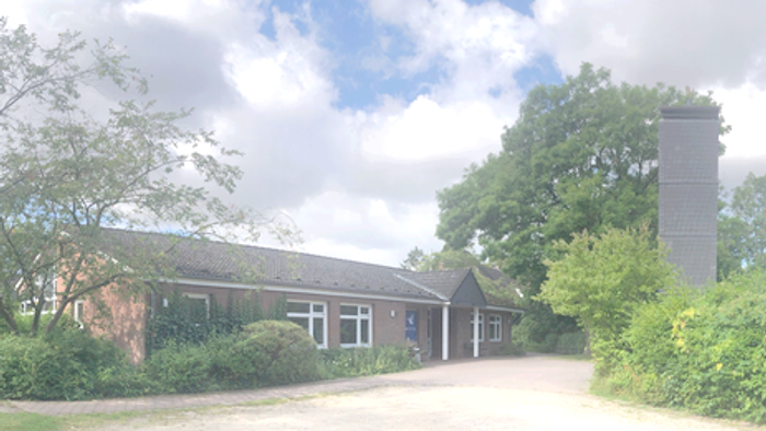 Ländlicher Gebäudekomplex, umgeben von Grünflächen, mit einem hohen Schornstein und klarem Himmel im Hintergrund.