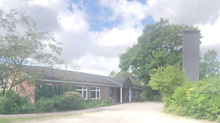 Ländlicher Gebäudekomplex, umgeben von Bäumen und Grünflächen mit einem hohen Schornstein und einem gepflasterten Weg.