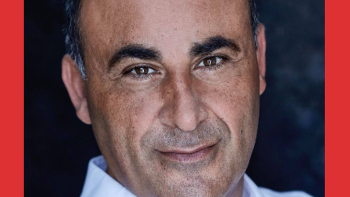 En mand med mørk hår og en hvid skjorte smiler til kameraet.