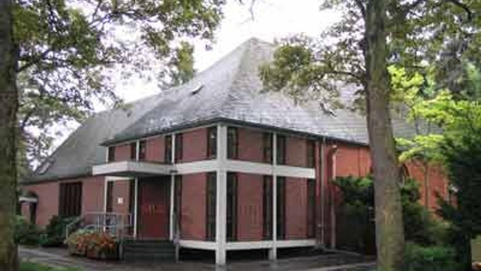 Das Bild zeigt ein zweistöckiges Haus mit rotem Dach und weißen Fensterläden, umgeben von Bäumen.