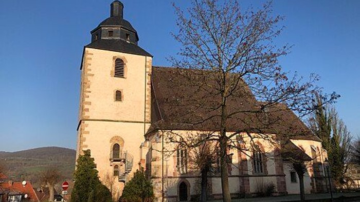 Kirche mit hohem Turm und Kreuz, umgeben von Bäumen und Gebäuden.