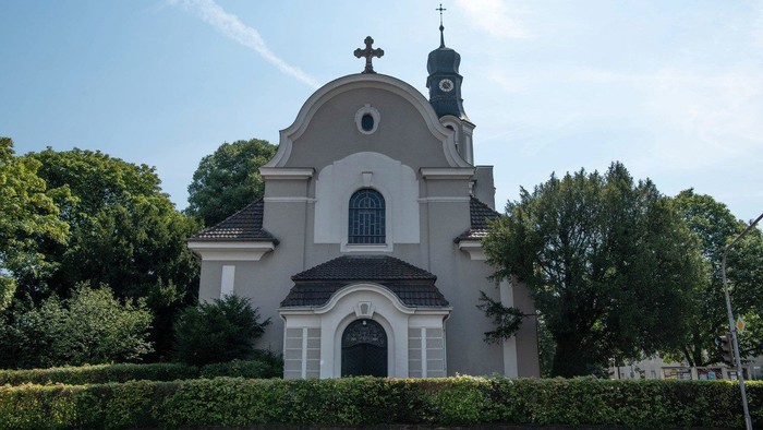 Kleine Kirche mit Kuppel und Kreuz auf dem Dach, umgeben von Bäumen und Büschen