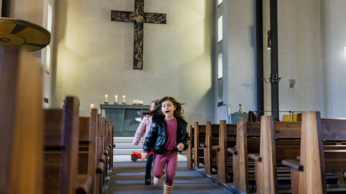 Ein junges Mädchen läuft den Mittelgang einer Kirche mit hölzernen Kirchenbänken und einem großen Kreuz an der Wand entlang.