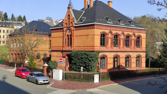 Ein großes, oranges Gebäude mit spitzen Dächern und einem gepflegten Garten, vor dem zwei Autos geparkt sind.
