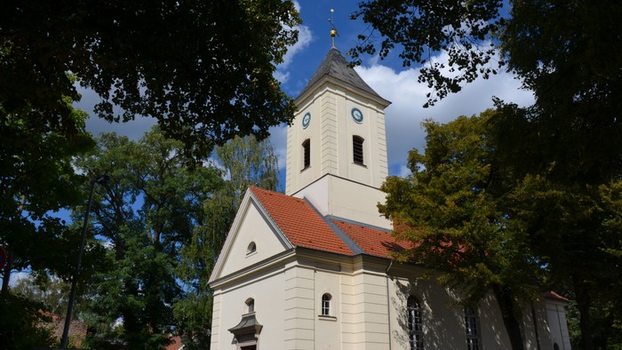 Weiße Kirche mit hohem Turm und roter Dachziegeln, umgeben von Bäumen