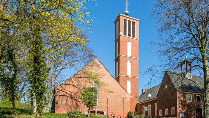 Kirchturm aus rotem Backstein mit Kreuz, umgeben von Bäumen und grünem Gras