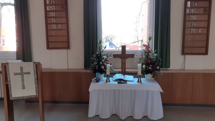 Ein Tisch mit weißer Tischdecke und Blumen in einem Raum mit zwei Kreuzen und Fenstern