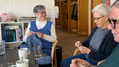 Tre voksne sidder og strikker og snakker omkring et bord med te og garn.